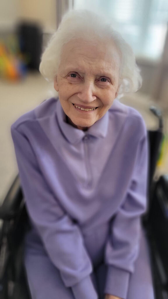 An older woman, Marilyn, is smiling and wearing a soft purple outfit.
