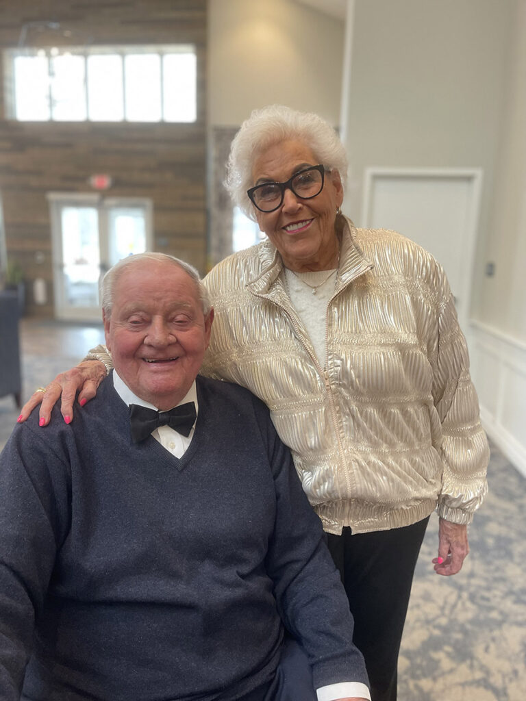 Elderly couple: woman in black glasses and a shimmering cream jacket, standing by a man in a blue v-neck sweater, collared shirt, and bow tie, sitting down.