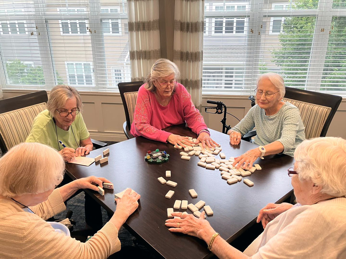 A group of senior living ladies sitting at a table having fun playing dominoes together.
