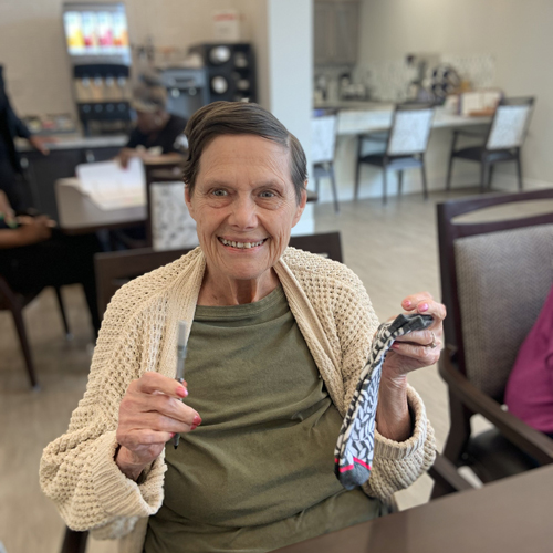 Senior woman wearing a green shirt and beige cardigan, sitting at a table in a senior living community, smiling and holding a pair of patterned socks and a sewing tool.