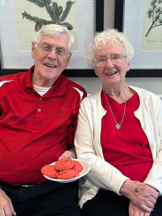 A senior couple dressed in red enjoys some cookies together, sharing a joyful moment in the community.