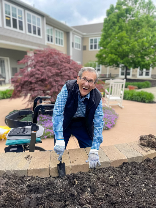 A senior resident enjoys gardening in the community garden. He is smiling while working with the soil and planting flowers.