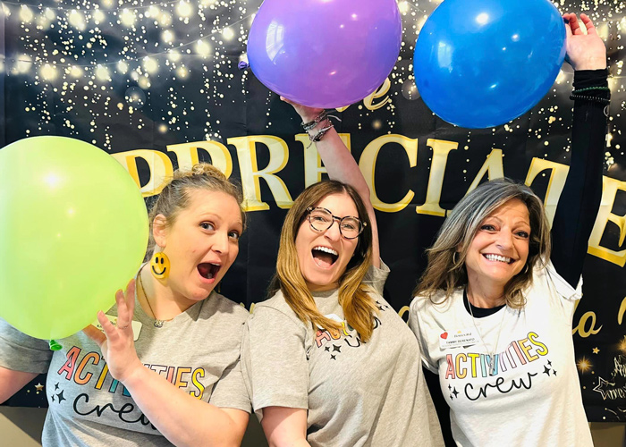 Three enthusiastic senior living team members from the Activities Crew hold colorful balloons and smile in front of an 'Appreciation' banner during a lively community event.