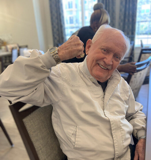 Smiling senior resident in a white jacket raises his arm in a playful flexing gesture while seated in a senior living dining area, exuding confidence and joy.
