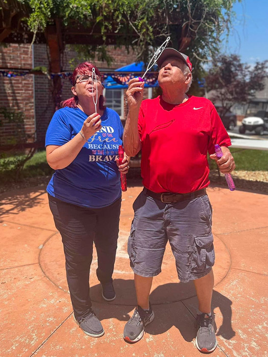 Senior resident and team member enjoy a sunny day blowing bubbles together outdoors, dressed in patriotic red and blue attire, celebrating a festive event with smiles and fun.