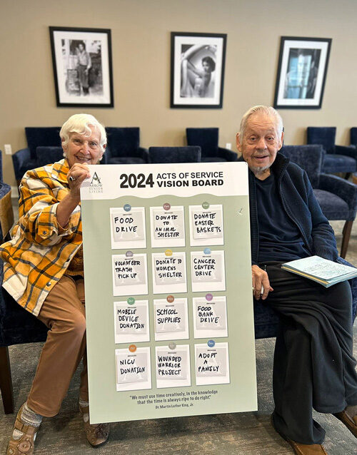 Two senior residents proudly display a large '2024 Acts of Service Vision Board' featuring various charitable activities, including food drives, donations, and volunteer work.