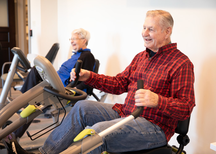 Two senior residents, a man in a red plaid shirt and a woman in a blue jacket, smile while exercising on recumbent bikes in a bright and welcoming fitness room.