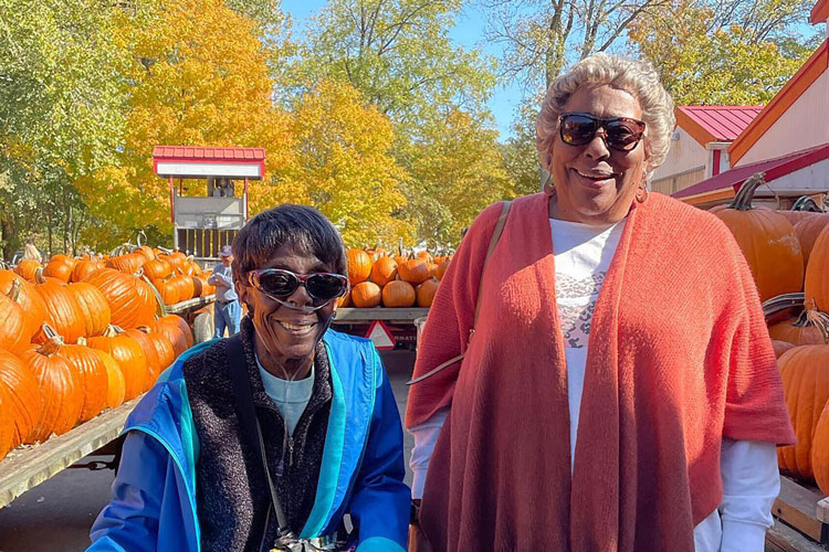 Two smiling senior residents wearing sunglasses enjoy a sunny autumn day at a pumpkin patch, surrounded by pumpkins and colorful fall foliage, during an outdoor outing.