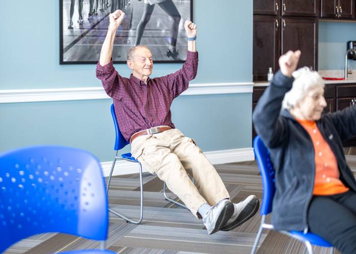 A senior resident sits in a blue chair, lifting his legs and raising his arms enthusiastically during a group exercise session in a well-lit room with blue chairs and a motivational poster.