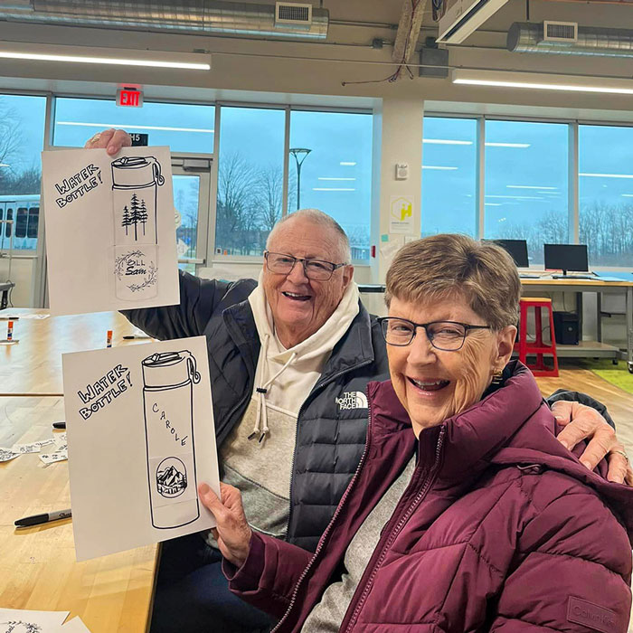 Smiling senior resident couple proudly display their custom water bottle designs on paper during an art activity at Lakeland Community College, enjoying a creative and fun experience together.