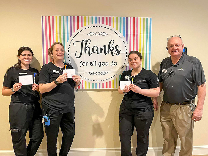Four team members stand in front of a 'Thanks for all you do' sign, holding appreciation gifts during an employee appreciation event at a senior living community.