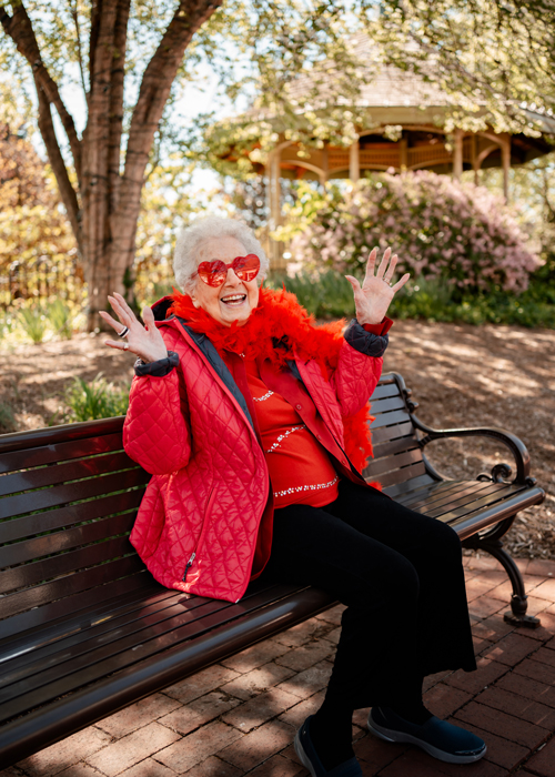 A joyful senior resident wearing red heart-shaped glasses and a feather boa, sitting on a park bench, smiling and waving, enjoying a beautiful day in a garden gazebo setting.