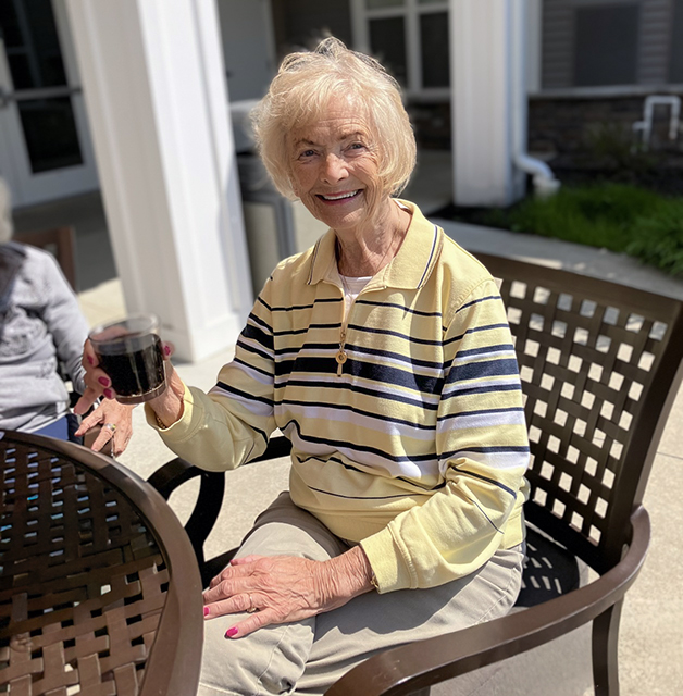 Senior woman in a striped shirt sits at a patio table outdoors, smiling while raising an icy soda.
