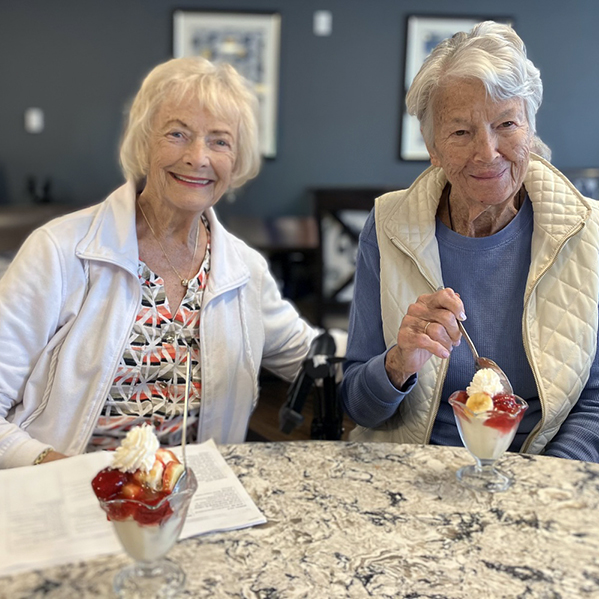 Two senior women sit at a marble table, smiling and eating ice cream sundaes.