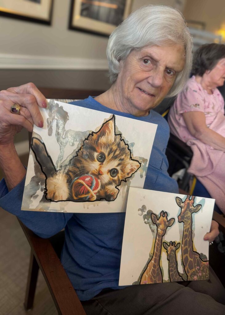 Senior woman proudly shows two finished art pieces, one of a kitten with yarn and another of three giraffes, during an engaging craft session in the community.