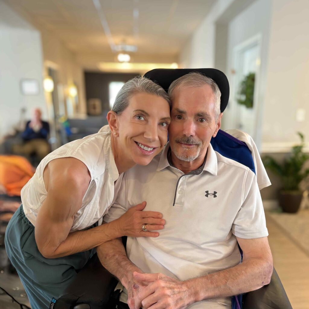 Woman smiles while leaning close to a man in a wheelchair, both wearing light-colored shirts, sharing a warm and supportive moment in a senior living community.