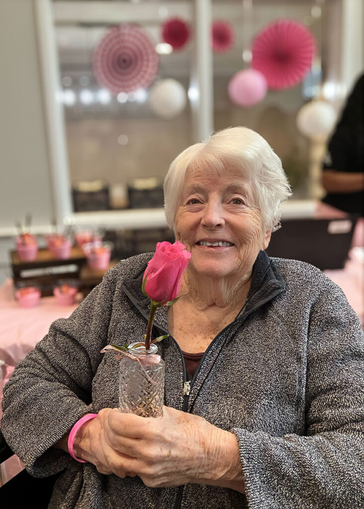A senior woman smiles, holding a glass vase with a pink rose.