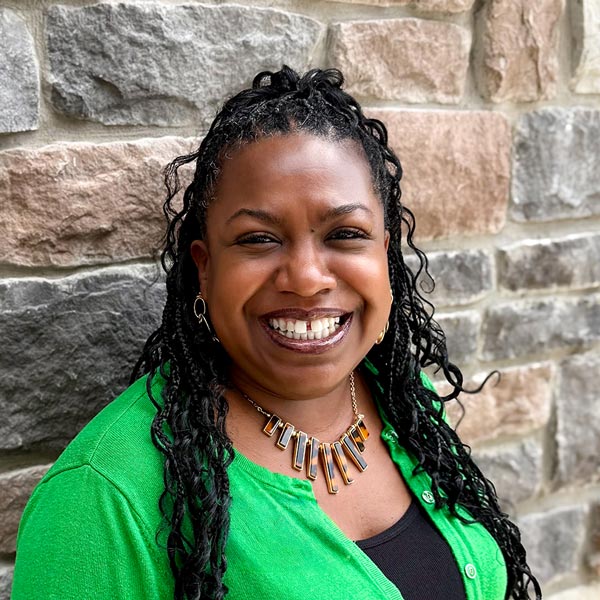 Cynthia Thompson, Business Office Director at Vitalia Westlake, smiling in a professional headshot. She has long braided hair, wears a green sweater over a black top with a statement necklace, and stands in front of a textured stone wall background.