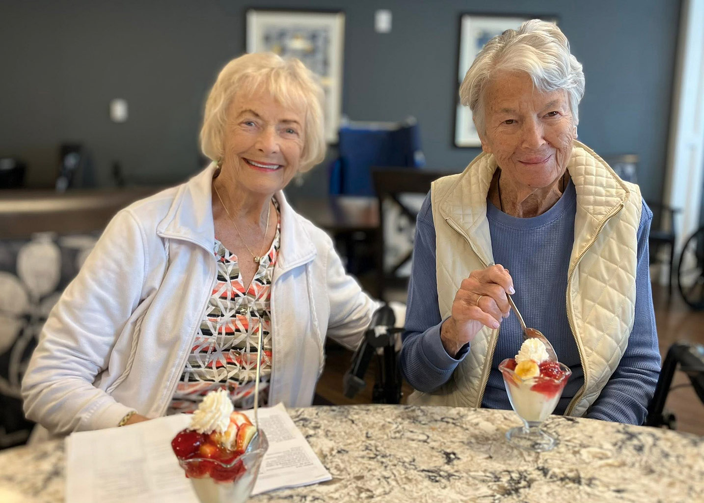 Two women sit together in a dining area, enjoying parfaits topped with strawberry sauce, fruit and whipped cream.