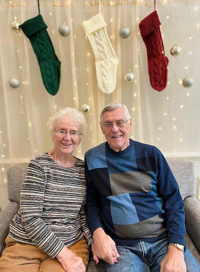 A senior man and woman smile in front of an elegant white winter backdrop and Christmas stockings.