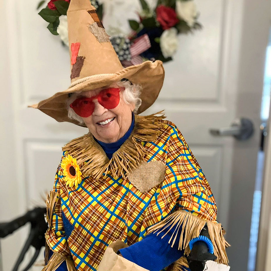 A senior woman smiles while wearing a scarecrow witch costume and novelty glasses.