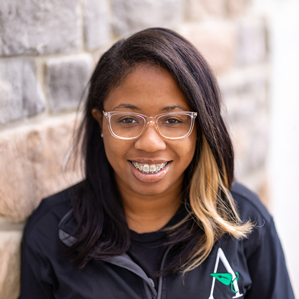 Charletta Richardson, Wellness Director at Vitalia Westlake, smiling in a professional headshot, wearing glasses and a black jacket, standing against a softly blurred stone wall background.