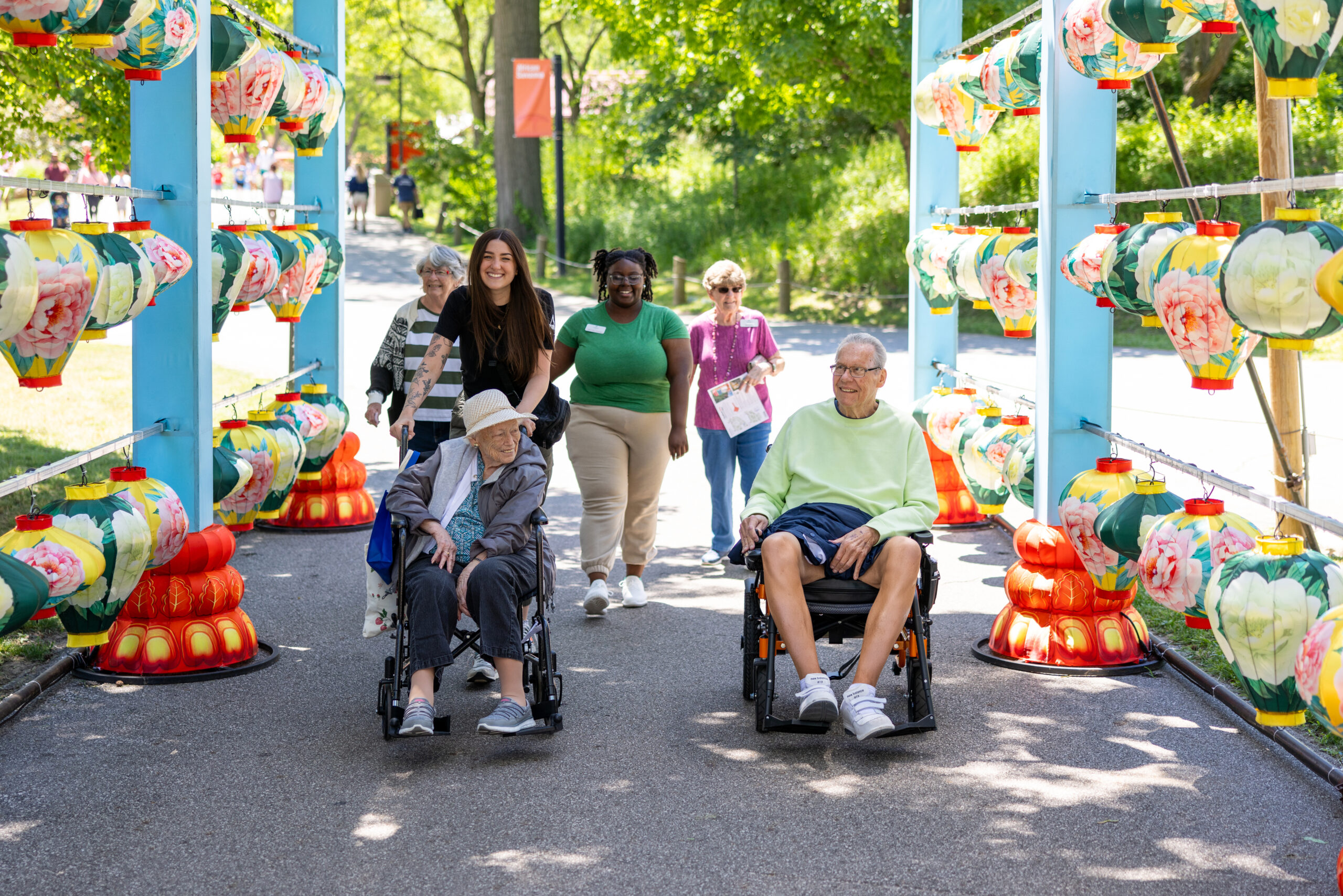 Group of senior residents, some in wheelchairs, enjoy a vibrant outdoor outing with colorful lanterns, accompanied by smiling caregivers on a sunny day in a scenic park.