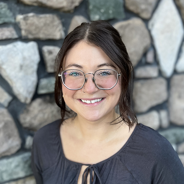 Jessie Cooper, Operations Coordinator at Vitalia Westlake, smiling while wearing glasses and a charcoal blouse, standing in front of a stone wall.