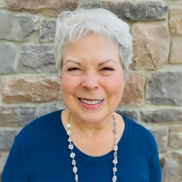 Marilynn “Ginny” Csider, Senior Living Coordinator at Vitalia Westlake, smiling warmly while wearing a blue top and necklace, standing in front of a stone wall background.