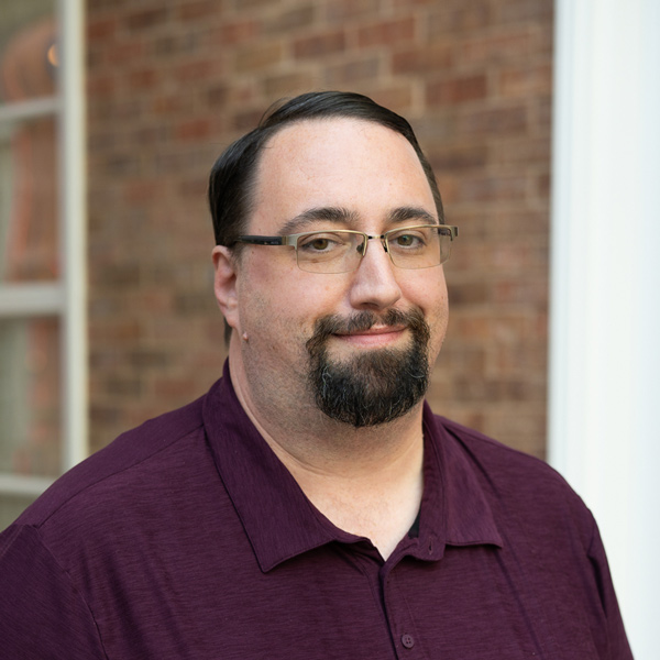 David Tinics, Plant Operations Director at Vitalia Westlake, smiling in a professional headshot, wearing glasses and a maroon polo shirt, with a softly blurred brick and column background.