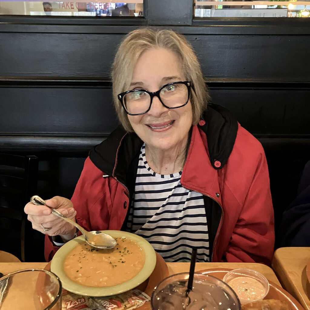 A woman wearing a red jacket smiles at a local eatery, enjoying a bowl of rich soup.