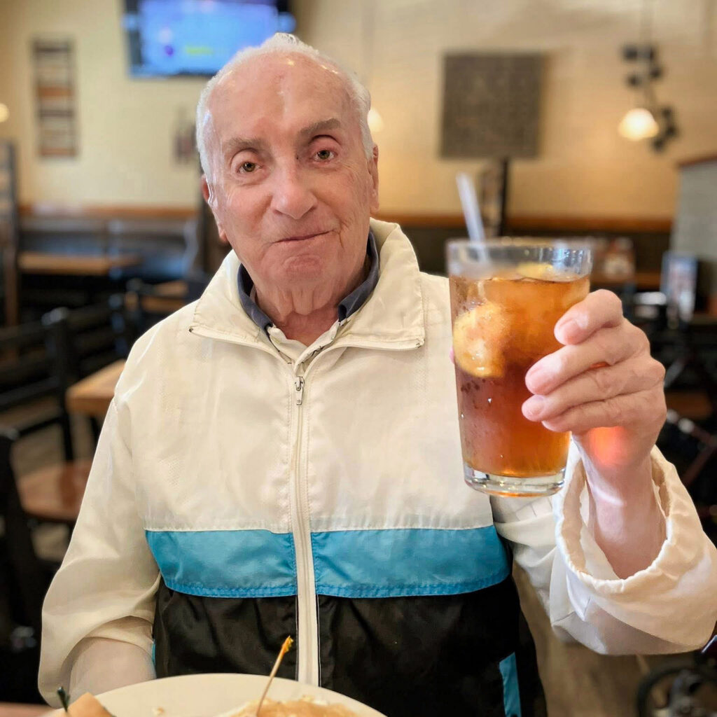 A senior man smiles in a relaxed manner, raising a glass of cold iced tea with lemon aloft.
