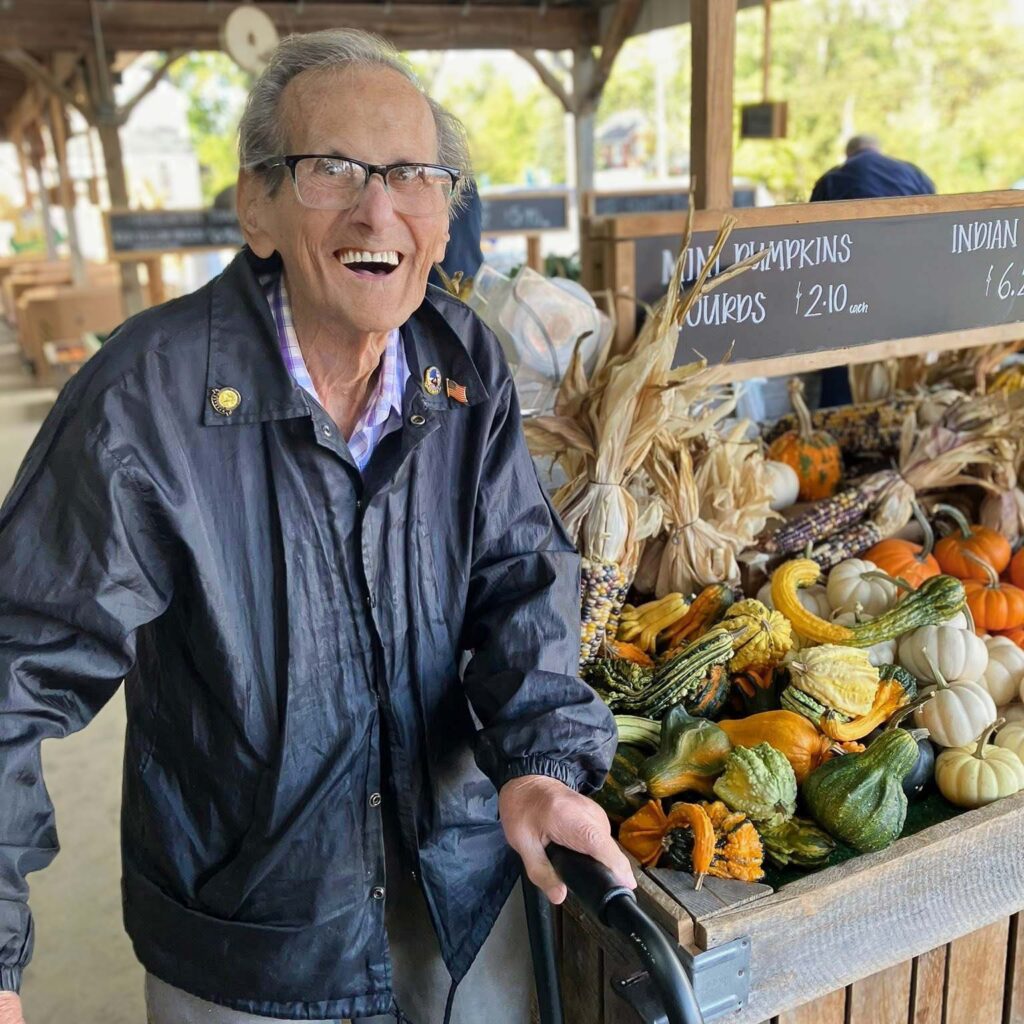 A senior man smiles delightedly at a local market, surrounded by heaping tables of gourds, corn, and seasonal products.