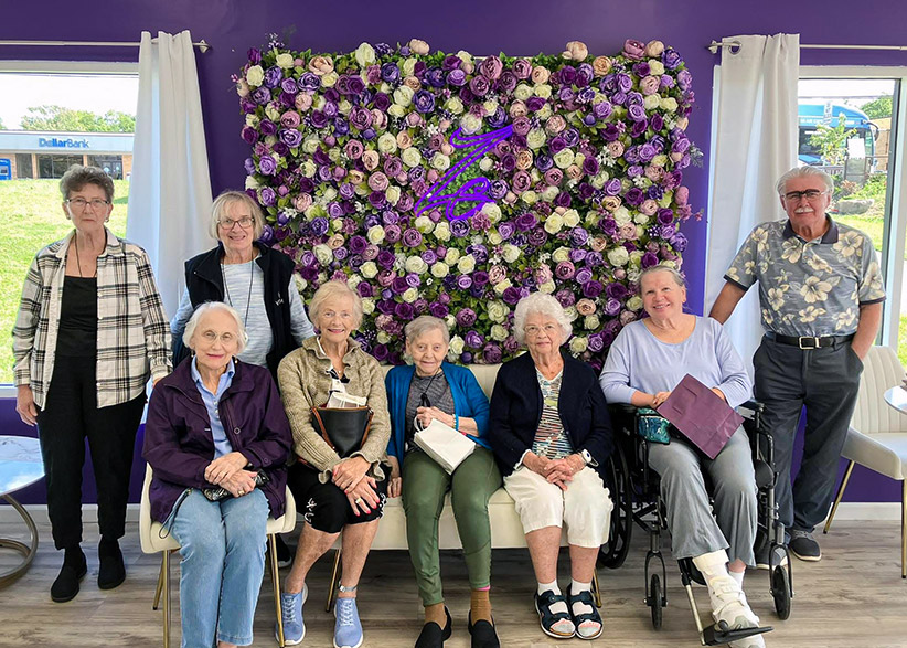 A group of senior residents from Vitalia Westlake pose for a photo at a local business, smiling in front of a purple wall featuring a large backdrop made of many purple, pink, and cream colored roses.