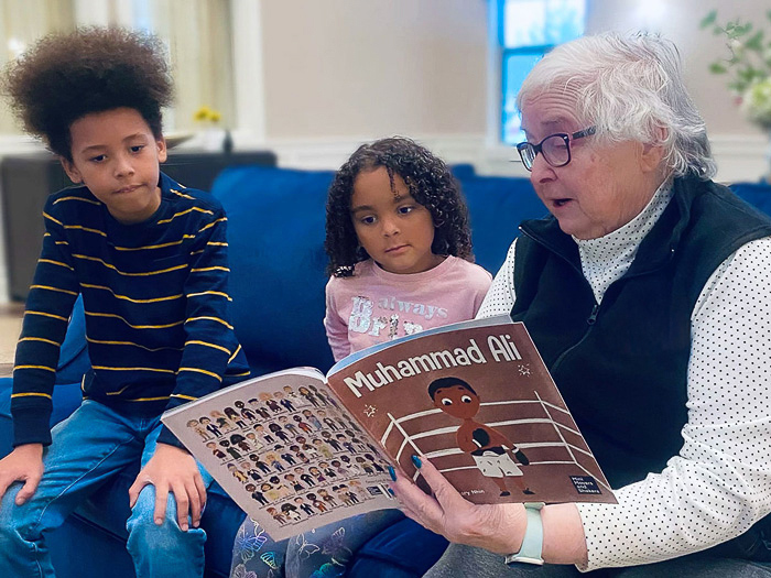 A senior resident reads a 'Muhammad Ali' children's book to two attentive young children, sitting together on a couch.