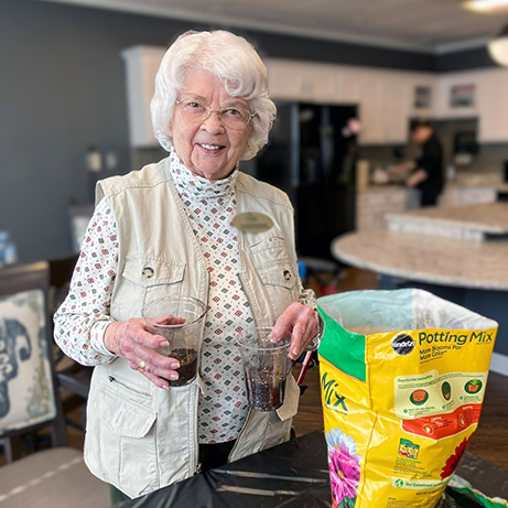 A senior woman wearing a gold Resident Ambassador badge smiles next to a bag of potting soil, cups in each hand, while working on a gardening project.