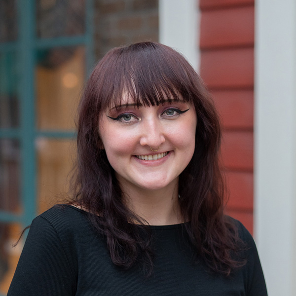 Ash Koenig, Arrow Memory Care Director providing Interim Executive Director support at Vitalia Westlake, smiling in a professional headshot with dark auburn hair, bangs, a black top, and teal window details in the background.